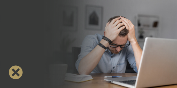 A man with glasses on sitting at a desk with a laptop in front of him. His head is in his hands to show that he is stressed to demonstrate that he has made a mistake. 
